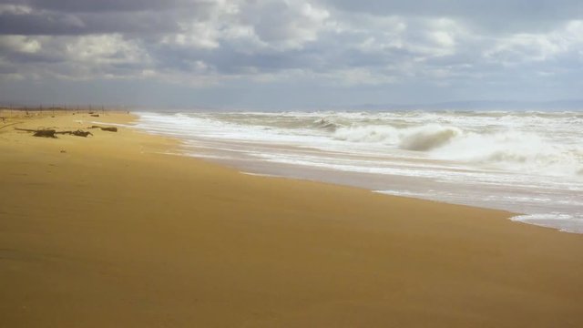 A sandy beach with a fence in the distance and a stormy sea with waves under a cloudy sky, a shadow from a cloud rushes to the beach