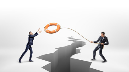 A businessman throws a lifeline with an orange life buoy over a large earthquake crack to another businessman.