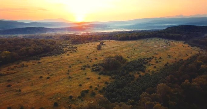 Flying backwards above the green leafy forest with field in the middle, hills covered with fog and red sun going down. Russia. The Far East