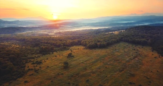 Flying above the green leafy forest with field in the middle, hills covered with fog and red sun going down. Russia. The Far East