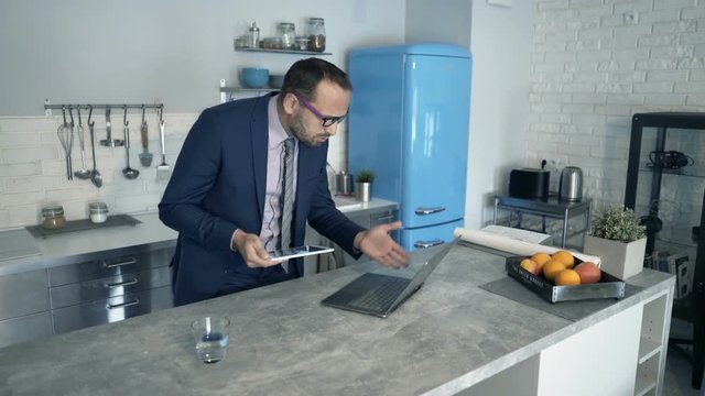 Unhappy businessman with laptop and tablet in kitchen at home
