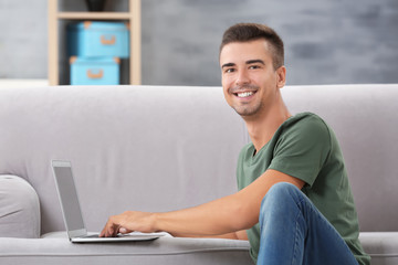 Young man with modern laptop near sofa at home