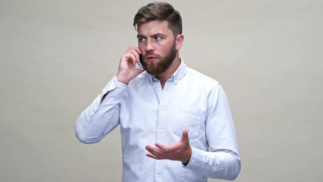 Worried bearded man in shirt talking by smartphone over gray background