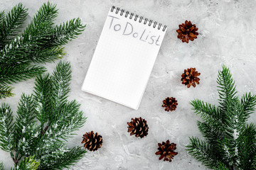To do list for winter shopping at notebook near spruce branch and pinecones on grey stone background top view