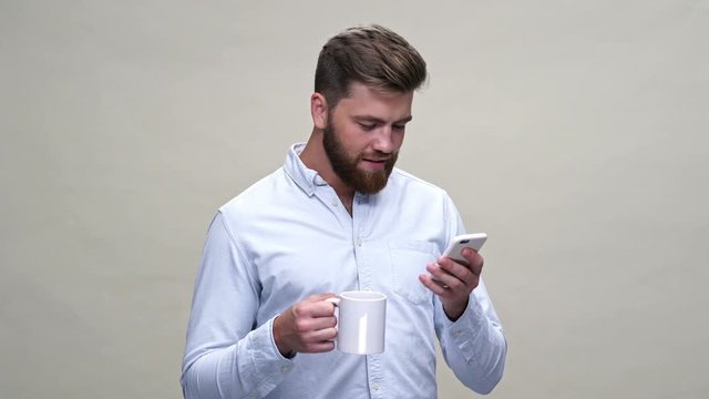 Surprised bearded man in shirt using smartphone while holding cup of tea over gray background