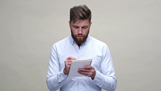 Concentrated pensive bearded man in shirt writing something in notebook over gray background