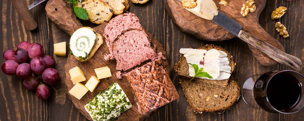 Assorted cheeses on wooden boards plate, grapes, bread wine and pate on dark wooden background, top view, flat lay, banner.
