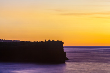 Silhouette of a cliff against the background of an orange sunset. Sea waves break on the rocks. Sunset on Jimbaran, South Kuta, Bali, Indonesia.