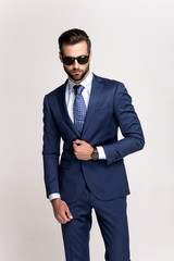 So stylish. Handsome young man in sunglasses adjusting his jacket and looking at camera while standing against white background.