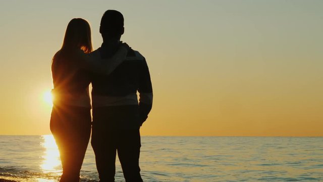 Silhouettes of a young couple hugging and admiring the sunset together by the sea. Back view