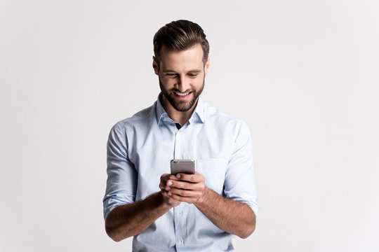 Great news! Handsome young man using his phone with smile while standing against white background.