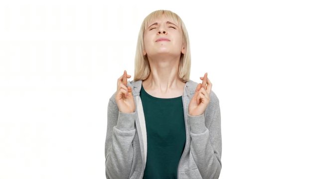 portrait of 20s blond woman wearing sweatshirt wishing for good luck looking up with closed eyes and keeping fingers crossed begging god please over white background closeup. Concept of emotions