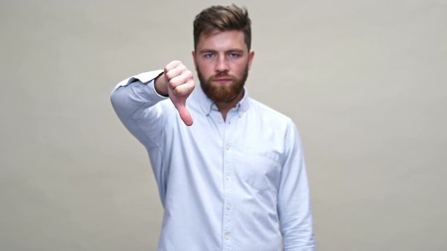Young bearded man in shirt showing thumb down at the camera over gray background