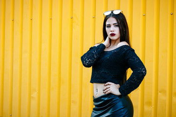 Gorgeous brunette girl with a beautiful make-up in fashionable clothes: a black jacket with a bare navel and leather skirt with shoes and sunglasses. Against the background of a yellow wall