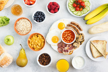 English breakfast fried eggs, sausages, bacon and mushrooms with selection of fruits and vegetables, breads and juice on the grey white table, top view, selective focus