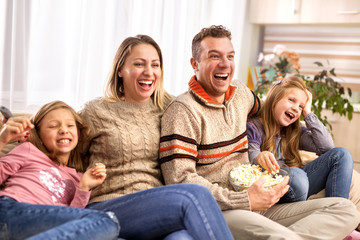 Beautiful young parents and their children are watching TV, eating popcorn and  sitting on couch at home