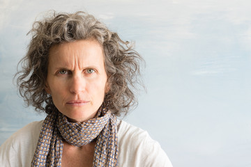 Middle aged woman with grey hair fowning and looking angry against blue background with copy space (selective focus) - obrazy, fototapety, plakaty