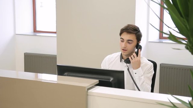 Male employee is talking on phone while sitting at desk at reception. Young man makes call with client, looking at black monitor of pc then hangs up. Promising manager dressed in white shirt works