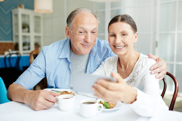 Fototapeta na wymiar Smiling senior couple making selfie by table in restaurant during dessert