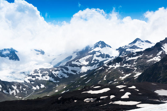 Fototapeta The beautiful mountain view in sunny day with blue sky and white clouds, black rocks and snow hills in Caucasus, in South Russia