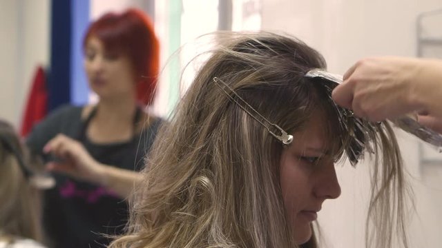 Hair coloring. Hairdresser Dye the Hair Dye Blonde Girl Sitting in a Beauty Salon, Barber Brush Hair Holds Paint, Apply the Foil and Wrap the Hair, a Man in a Purple Shirt With a Black Apron, Mirror