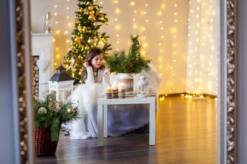 A brunette gilr in front of fur-tree and fireplace with candles and gifts. A surprised girl. A girl dreaming. New year's eve. Christmas eve. Cozy holiday at the fur-tree with lights and gold decor.
