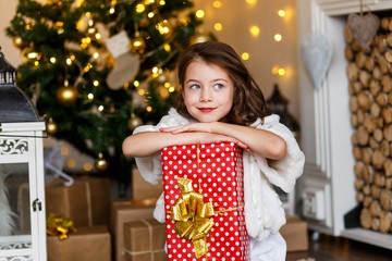 A brunette gilr in front of fur-tree and fireplace with candles and gifts. A surprised girl. A girl dreaming. New year's eve. Christmas eve. Cozy holiday at the fur-tree with lights and gold decor.