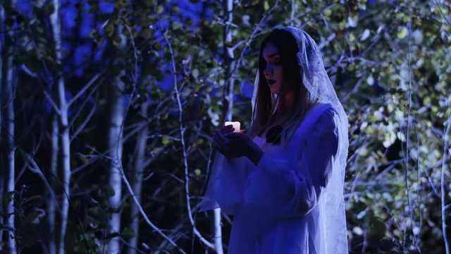 The young woman with skeleton make-up for Halloween in the white bride dress holding in the arms a burning wax candle in the dark forest at night. 4K