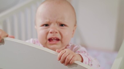 Baby's standing in a white crib at home and crying. Close-up shot.