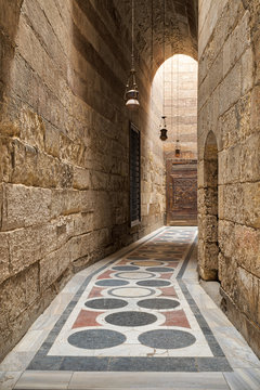 Arched corridor leading to the courtyard of Sultan Barquq mosque with stone bricks wall, ornate colored marble floor and wooden door, Old Cairo, Egypt