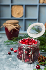 Fresh juicy cranberries in a glass jar on a blue marble background with spruce branches and cones with copy space