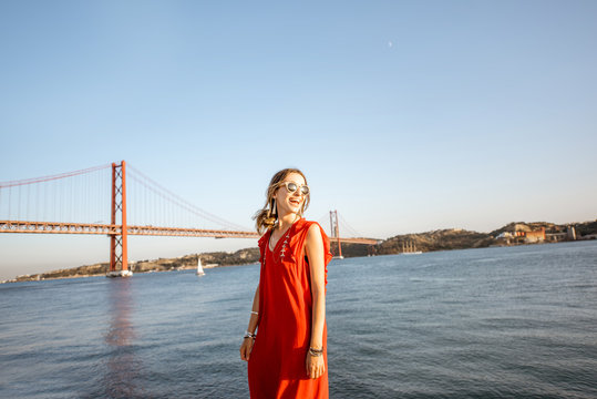 Lifestyle portrait of a woman in red dress walking on the riverside with beautiful iron bridge on the background in Lisbon city, Porugal