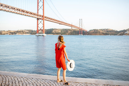 Woman in red dress enjoying landscape view on the famous iron bridge standing back on the riverside in Lisbon city.