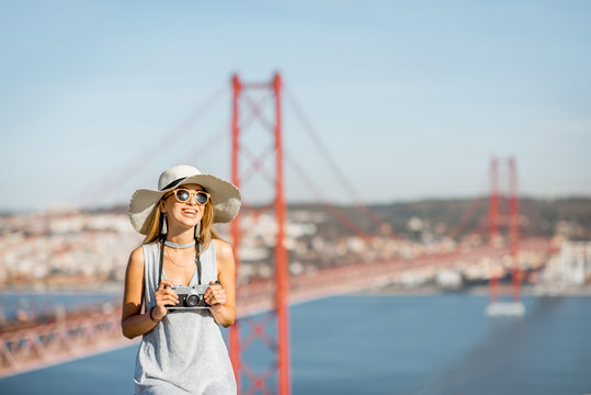 Portrait of a young woman tourist with photo camera sitting on the landscape view background with beautiful iron bridge in Lisbon city, Portugal