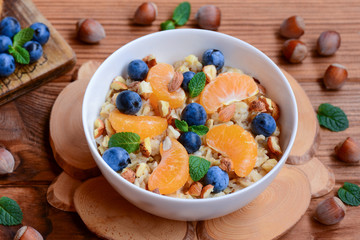 Delicious and diet porridge with fruits, berries and nuts. Classic oatmeal with fresh mandarins, blueberries, hazelnuts and mint in a white bowl. Wooden table. Porridge breakfast recipe. Rustic style