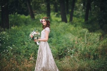Fototapeta na wymiar An elegant bride with a wreath on her head and a light cream wedding dress is standing in a summer forest. Walk on the nature.