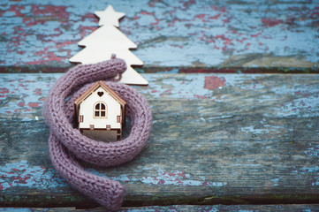 Toy house is wrapped in a warm scarf, it's snowing. in the background a fur-tree.