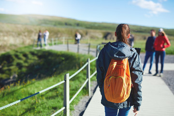 Naklejka premium back view of young woman hiker hiking in beautiful mountains. female hiker with small orange packpack black lava rocks background. excursion to the national park Thingvellir in Iceland
