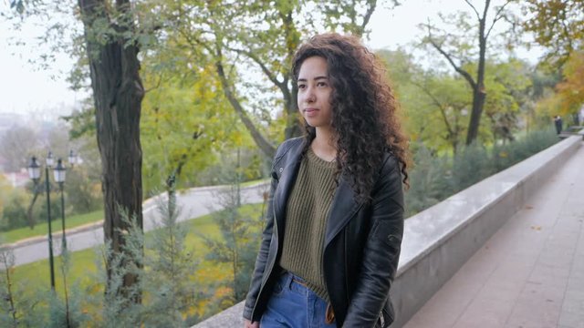 Portrait of happy young latin woman with beautiful curly hair walking in the street, slow motion. Hispanic girl in black leather jacket smiling and goes to camera