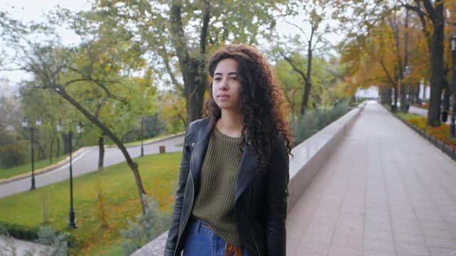 Portrait of happy young afro woman with beautiful curly hair walking in the street, slow motion. Hispanic girl in black leather jacket smiling and goes to camera