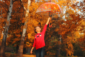 A girl in a red sweater is standing in an autumn park with an umbrella in her hands