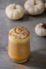 Glass of spicy pumpkin latte with whipped cream and cinnamon standing on gray kitchen table with decorative white pumpkins. Coffee beans and spices above. Atmospheric breakfast