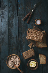 Variety loaves of sliced homemade rye bread whole grain and seeds for breakfast with olive wood bowls of butter, salt, liver paste over old dark wooden background. Top view, space. Toned image
