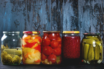 Variety glass jars of homemade pickled or fermented vegetables and jams in row with old dark blue...