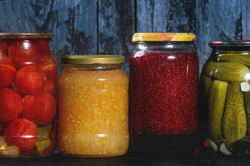 Variety glass jars of homemade pickled or fermented vegetables and jams in row with old dark blue wooden plank background. Seasonal preserves.