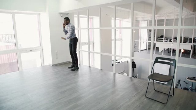 Young afroamerican man is talking on phone, walking around modern office. African male makes important call, using smartphone and gesticulates with hands actively, goes around light room. Successful