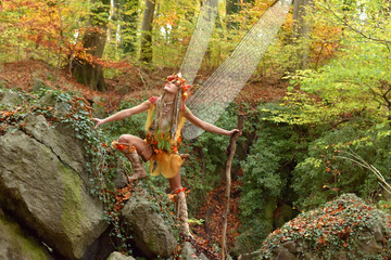 A beautiful young woman dresses up as an Autumn fairy.   She wears a fairy costume with wings on her back. She poses  for the camera in an enchanted Autumn forest.