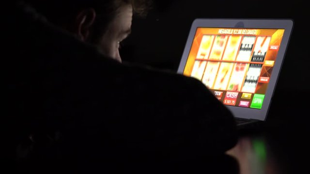 Gambling addicted man in front of online casino slot machine on laptop computer at night - loosing his money. Dramatic low light grain shot.