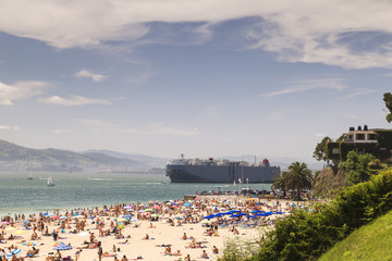 Large cargo ship leaving the harbor near a crowded beach
