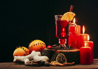 Glass of christmas hot mulled wine on wooden table with species ,oranges red candles against black background.Copy space